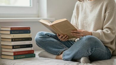 Pessoa sentada confortavelmente lendo um livro em um ambiente aconchegante, com luz natural e livros empilhados ao lado.